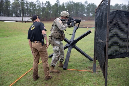 Man in USAMU shooting uniform instructing U.S. Army Soldier on outdoor firing range.