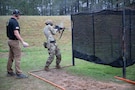 Man in USAMU shooting uniform instructing U.S. Army Soldier on outdoor firing range.