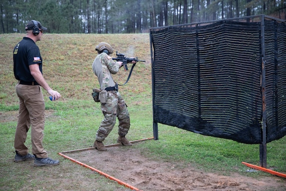 Man in USAMU shooting uniform instructing U.S. Army Soldier on outdoor firing range.