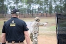 Man in U.S. Army uniform firing pistol on outdoor pistol range while USAMU team member observes.