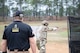 Man in U.S. Army uniform firing pistol on outdoor pistol range while USAMU team member observes.