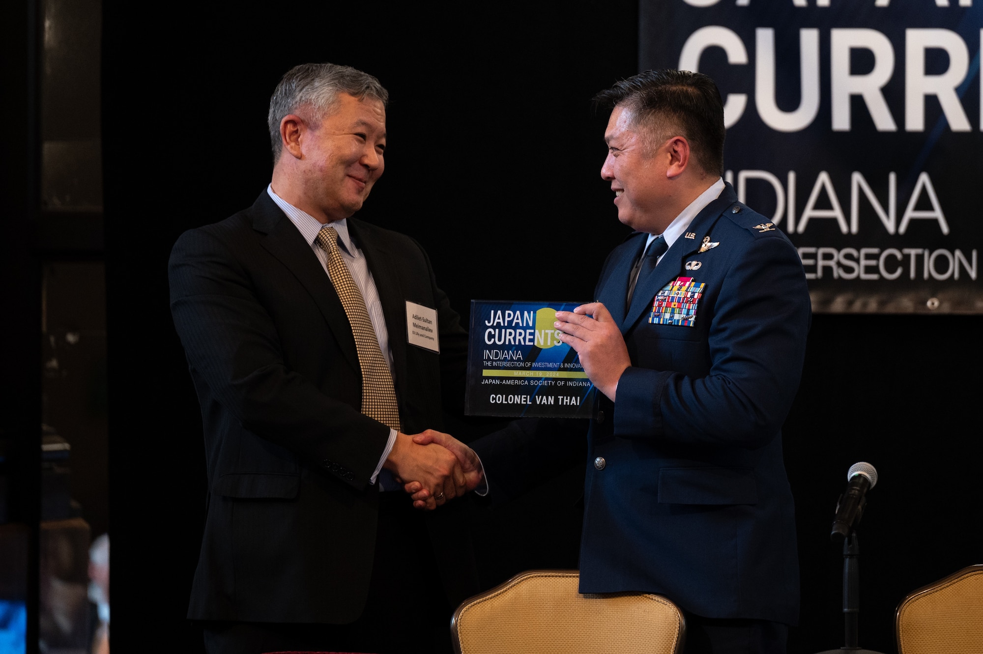A man in a suit shakes hands with a man in an Air Force service dress uniform. The man in the military uniform is holding a plaque that reads "JAPAN CURRENTS" "INDIANA" "COLONEL VAN THAI". Both men are smiling.