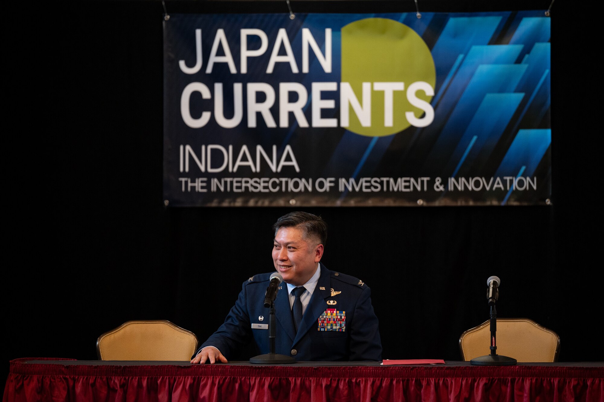 Col. Thai, in full Air Force service dress uniform, smiles while looking into a crowd. He is sitting down. On either side of him, two yellow chairs. A banner can be seen above his head that reads "JAPAN CURRENTS" "INDIANA" "THE INTERSECTION OF INVESTMENT & INNOVATION".