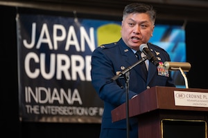 Col. Thai, in an Air Force service dress uniform, speaks into a microphone at a podium. In the background, a multi-color banner. His body blocks a portion of the banner, but "JAPAN CURR" can be read, as well as "INDIANA" and "THE INTERSECTION OF".