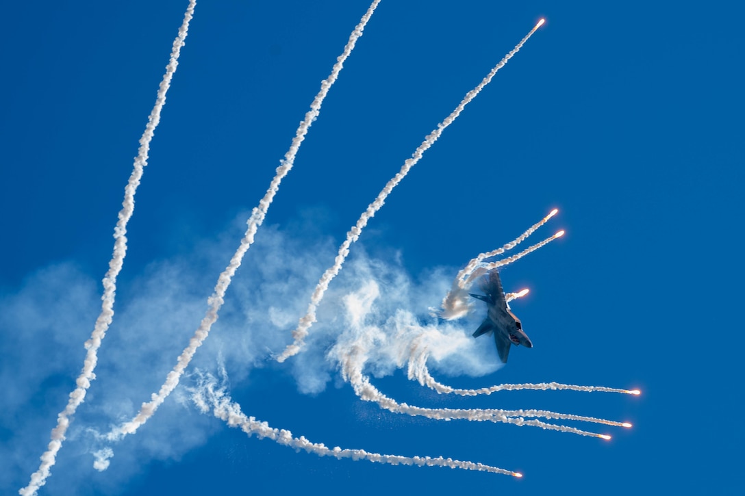 An aircraft releases rows of flares with smoke on either side while performing against a solid blue sky.
