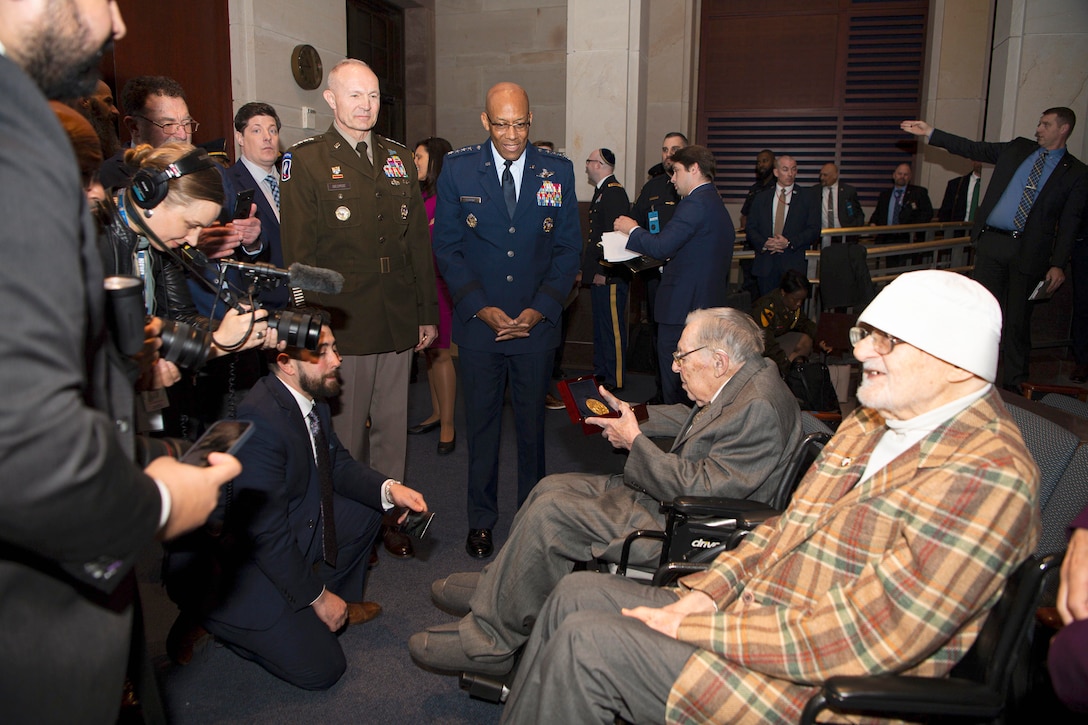 Members of the media film and interview seated veterans as two defense leaders stand to their right in a crowded room.