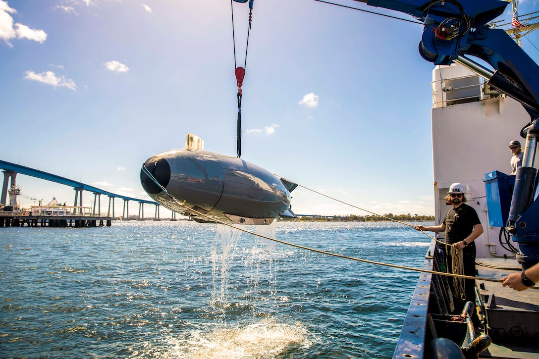 Mariners hold ropes attached to an unmanned vehicle above a body of water while standing on a boat with a bridge in the background.