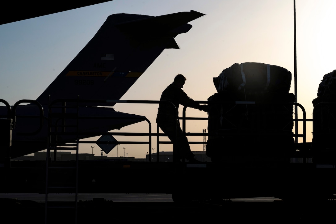 An airman pulls a pallet of supplies near the rear of an aircraft as seen in silhouette.