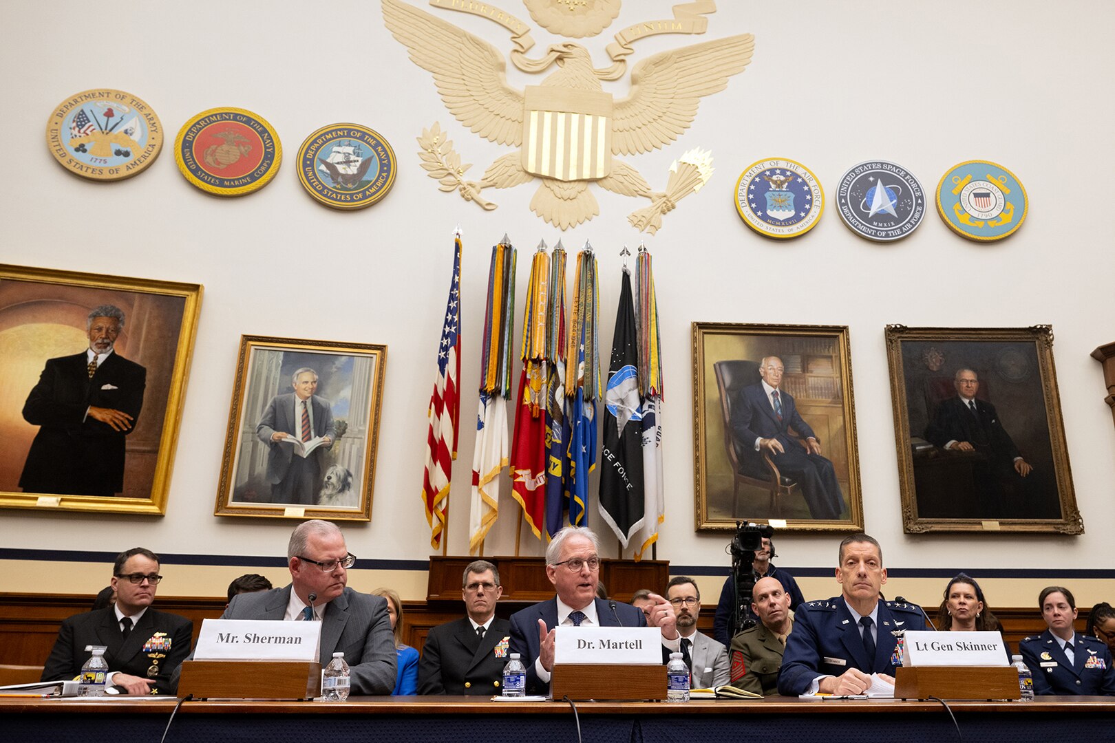 Two men in suits and one man in military uniform sit at a table inside a congressional hearing room.