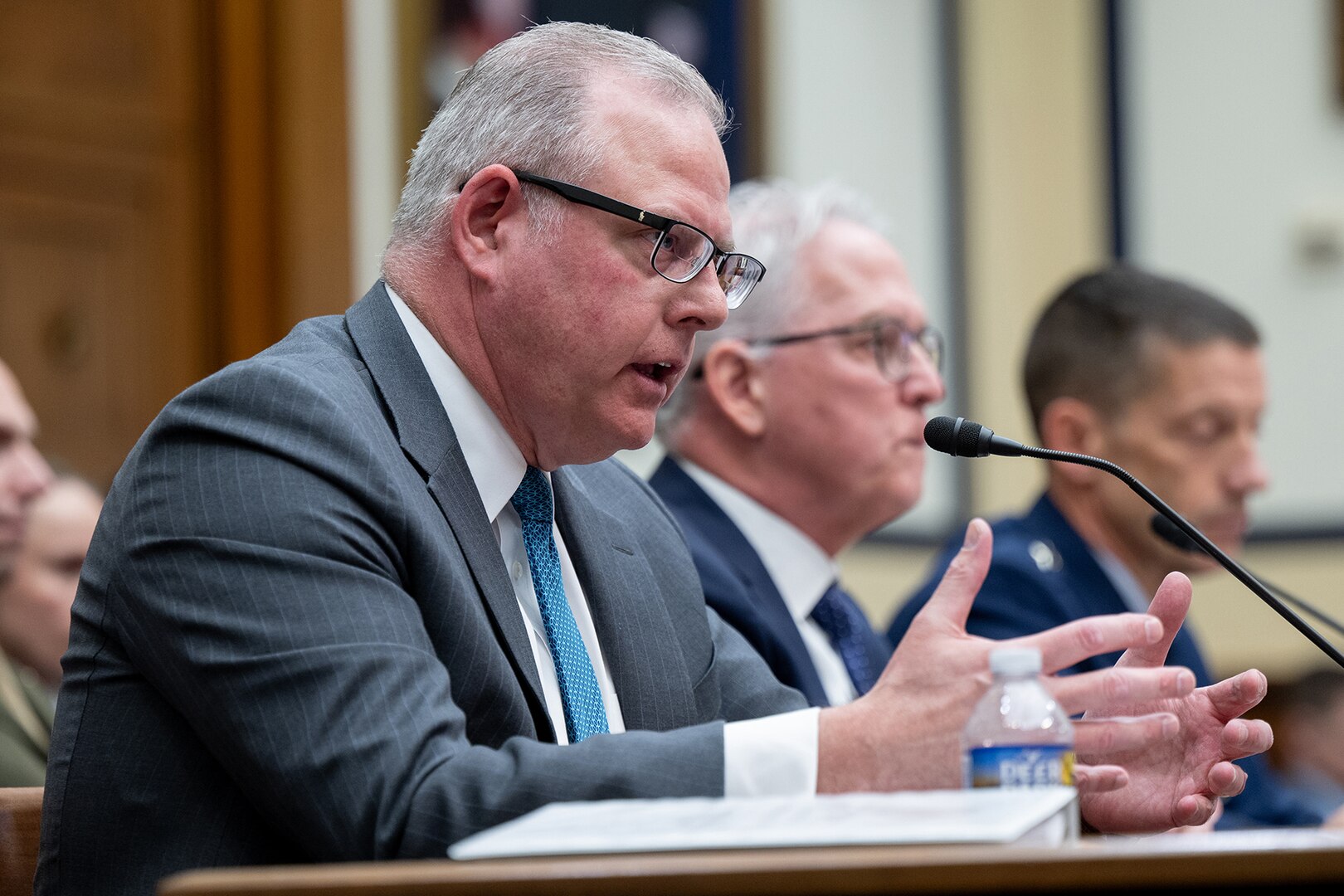 A man in a suit gestures with his hands while seated at a table inside a congressional hearing room.