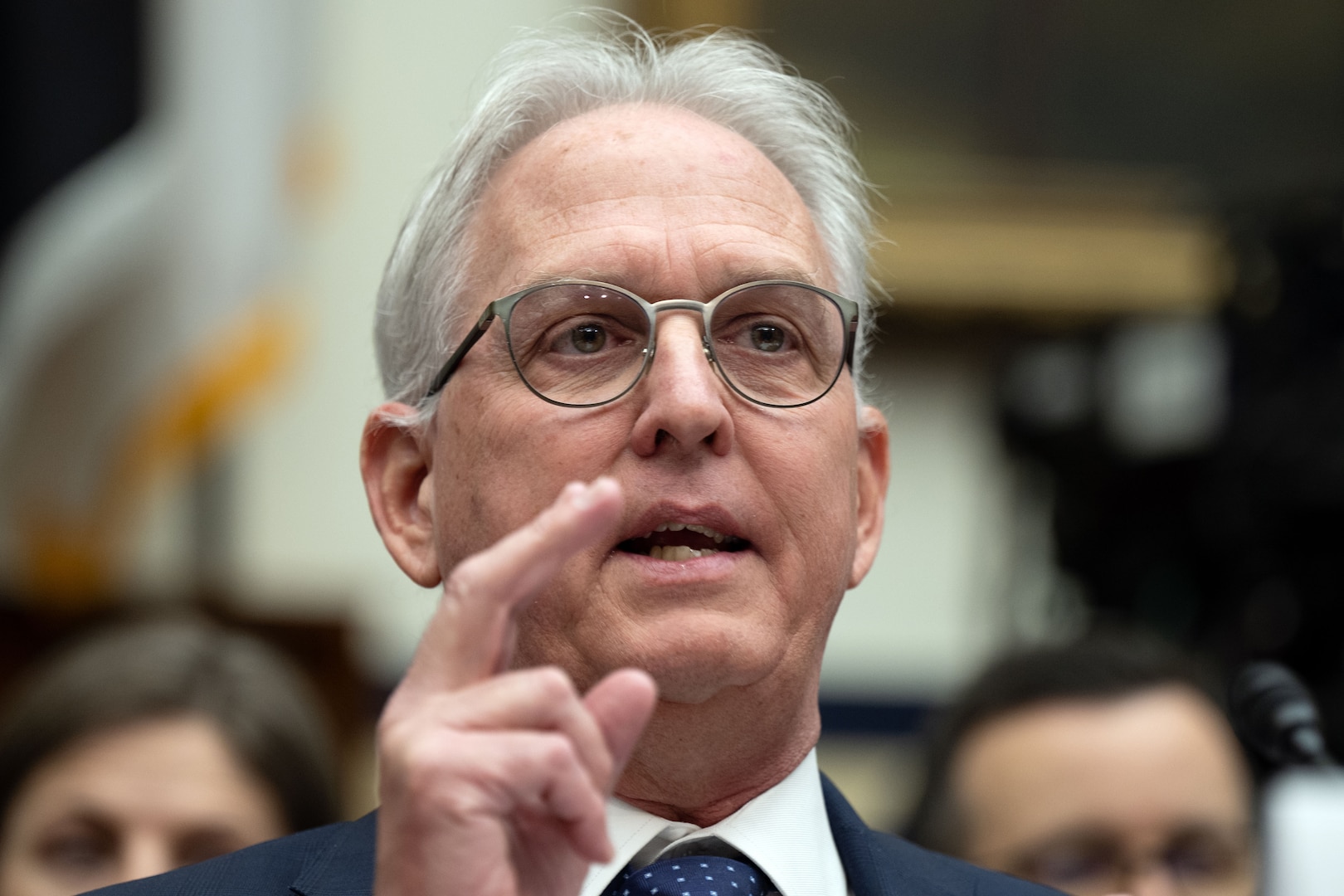 A man in a suit gestures with his hands while seated at a table inside a congressional hearing room.