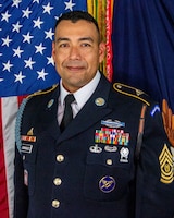 Army soldier with dark, short hair wearing Army dark green service uniform with tie, ribbons, badges and nametag. There is a US flag and another flag in the background.