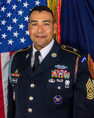 Army soldier with dark, short hair wearing Army dark green service uniform with tie, ribbons, badges and nametag. There is a US flag and another flag in the background.