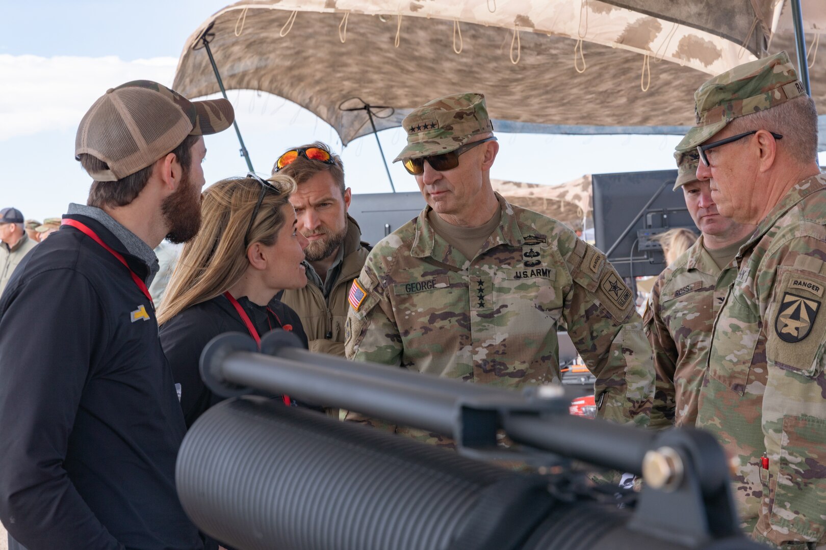 Two people in plain clothes talk to three soldiers in green camouflage with a large black cylindrical item in the foreground.
