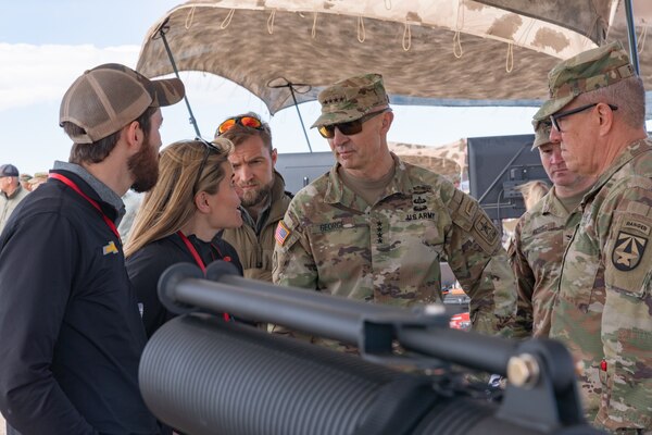 Two people in plain clothes talk to three soldiers in green camouflage with a large black cylindrical item in the foreground.