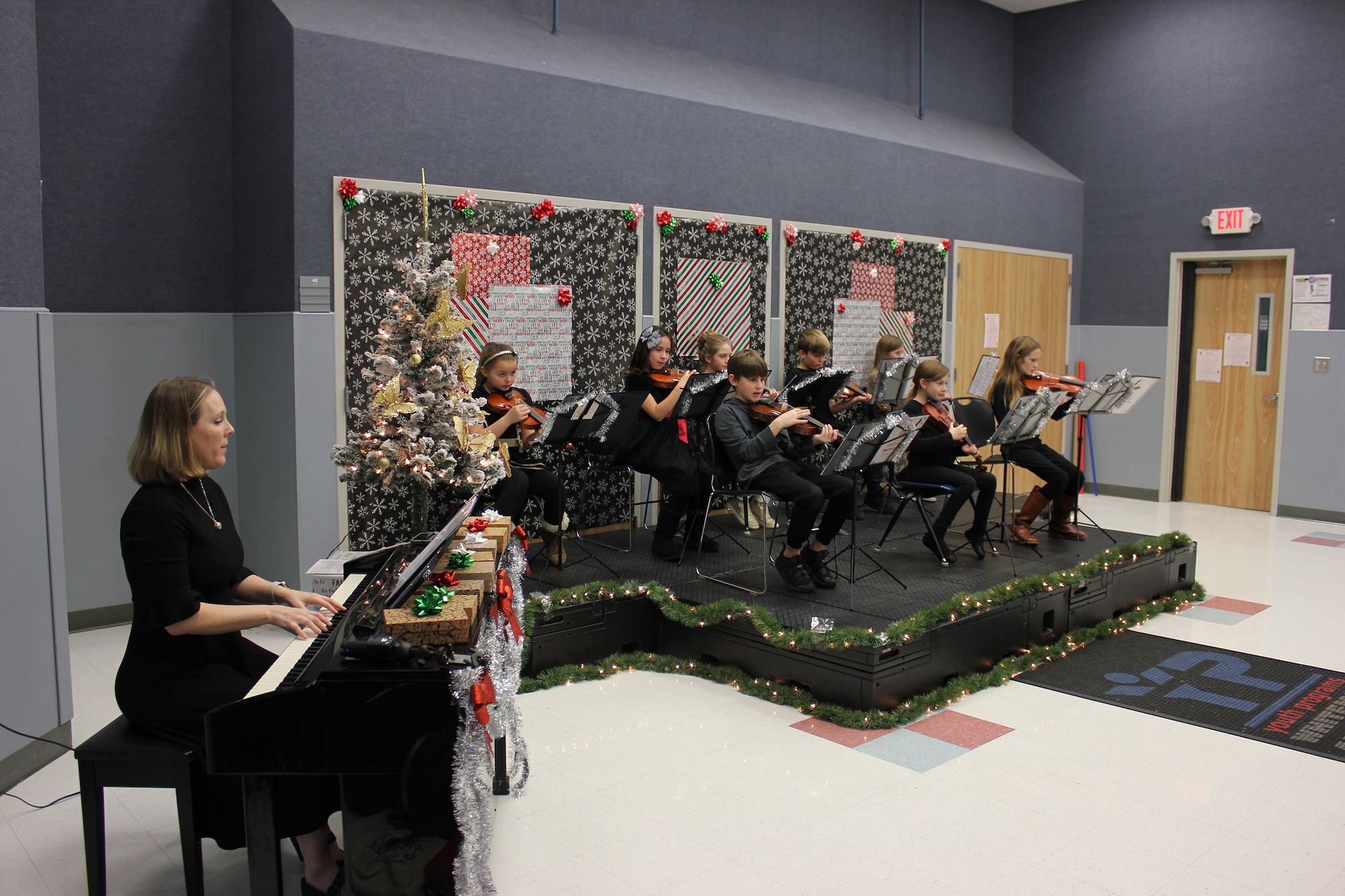 A children's orchestra performs songs on violin during a concert. An adult accompanies them on piano.