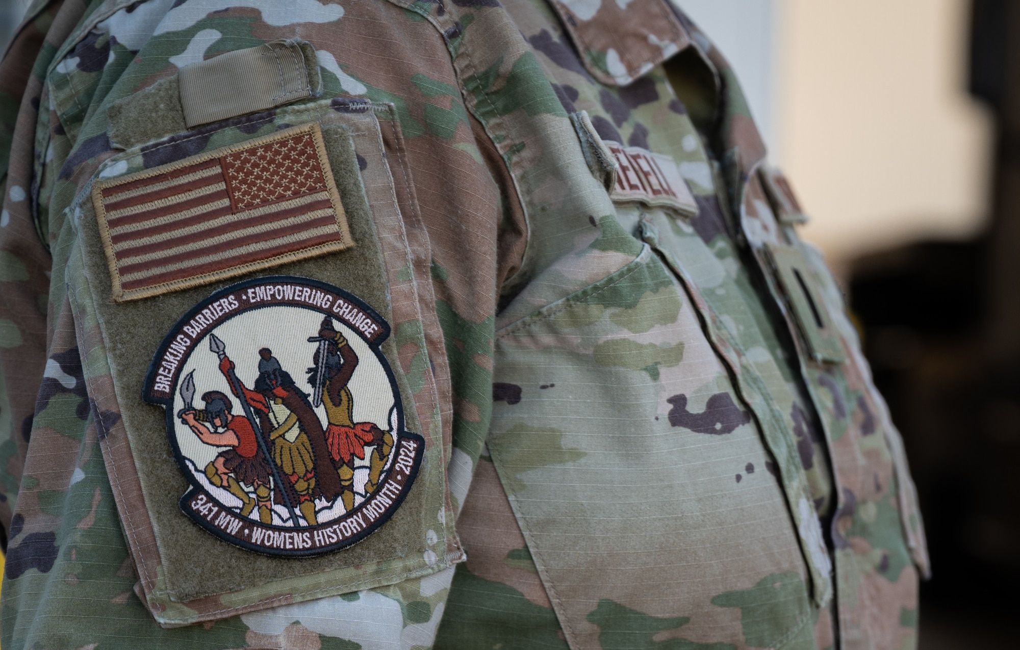 The side view of a person in military uniform is shown, focusing on their upper torso and right sleeve. On the sleeve is a circular velcro patch depicting three Spartan women charging into battle, surrounded by text that reads: "BREAKING BARRIERS - EMPOWERING CHANGE - 341 MW - WOMENS HISTORY MONTH - 2024"