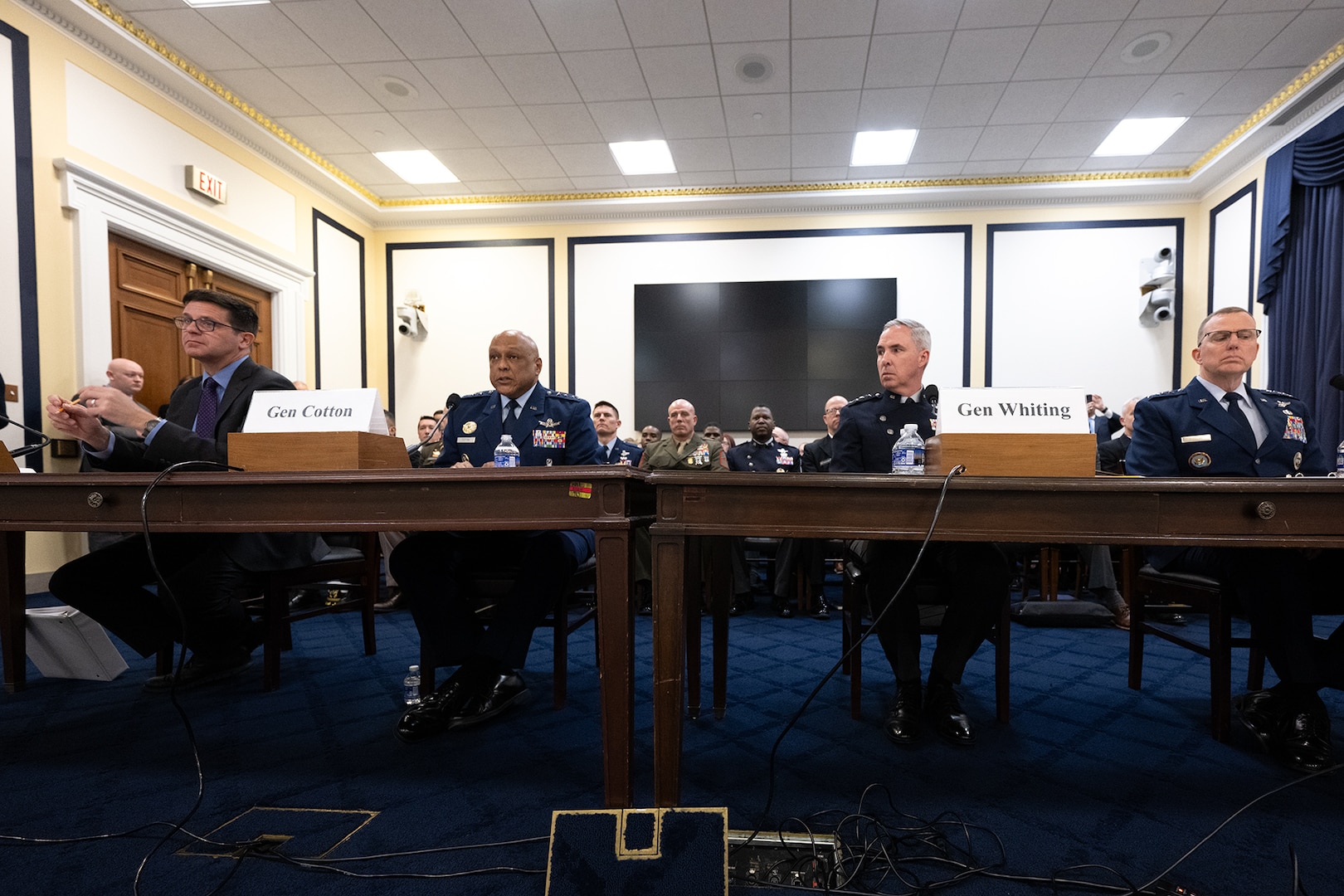 Four men sit near each other at two wooden tables. One wears a civilian suit, the other three wear military uniforms.