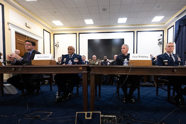 Four men sit near each other at two wooden tables. One wears a civilian suit, the other three wear military uniforms.