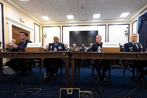 Four men sit near each other at two wooden tables. One wears a civilian suit, the other three wear military uniforms.