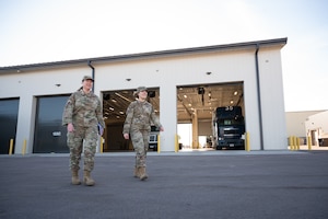 Two women in military uniform walk on grey concrete from the left side of frame toward the right.