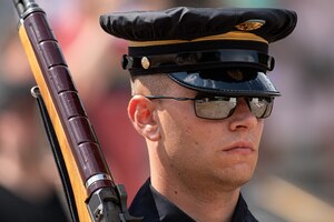 Closeup of soldier carrying rifle over right shoulder and looking away from the camera, wearing black sunglasses and a hat with the rain cover on it.