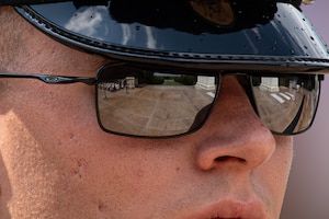 Closeup of a white man's face, wearing black sunglasses and a wheel hat with a glossy brim. There are some drops of water on the brim of the hat.