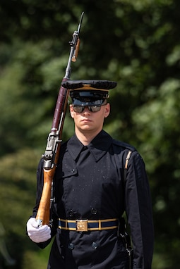 An Army soldier wearing a dark ceremonial uniform with a raincoat on is holding a large brown wood rifle on his right shoulder. He has a belt witha  gold belt buckle. Behind him are dark green trees.