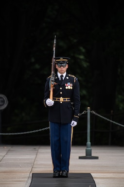 An Army soldier wearing a dark ceremonial uniform is standing at attention with his left hand at his side and his right hand holding a brown wooden rifle against his right shoulder. He is standing on a dark mat that is on a white marble plaza.