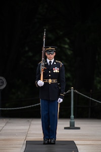 An Army soldier wearing a dark ceremonial uniform is standing at attention with his left hand at his side and his right hand holding a brown wooden rifle against his right shoulder. He is standing on a dark mat that is on a white marble plaza.