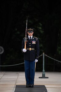 An Army soldier wearing a dark ceremonial uniform is standing at attention with his left hand at his side and his right hand holding a brown wooden rifle against his right shoulder. He is standing on a dark mat that is on a white marble plaza.