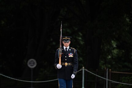 An Army soldier wearing a dark ceremonial uniform is standing still with a large brown wood colored rifle with bayonet affixed over his right shoulder.