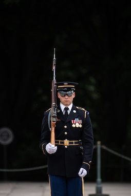 A Soldier is dressed in a dark ceremonial uniform, standing at attention while holding a brown wood rifle over his right shoulder. The background is very dark. There is a chain barrier in the background to keep people away from the area where the soldier is standing.