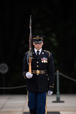 Army soldier in dark ceremonial uniform (dark jacket with gold buttons, gold belt buckle, white gloves, dark wheel hat with gold and yellow trim, and shiny medals on multicolored ribbons) and wearing sunglasses is facing the camera while either marching or standing.