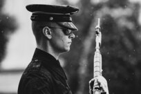 Black and white image of soldier facing the right of the picture, wearing dark ceremonial uniform with raincoat and rain cap on hat, wearing sunglasses, holding rifle in front of body with bayonet affixed. It is lightly raining.