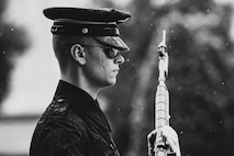 Black and white image of soldier facing the right of the picture, wearing dark ceremonial uniform with raincoat and rain cap on hat, wearing sunglasses, holding rifle in front of body with bayonet affixed. It is lightly raining.