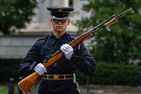 Army soldier wearing dark ceremonial uniform with raincoat and rain cap cover for his hat is carrying a brown wood rifle in both hands at a 45 degree angle in front of his body. The rifle has a bayonet affixed and he is marching or standing facing the cameraman. There are green trees and shrubs in the background