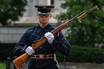 Army soldier wearing dark ceremonial uniform with raincoat and rain cap cover for his hat is carrying a brown wood rifle in both hands at a 45 degree angle in front of his body. The rifle has a bayonet affixed and he is marching or standing facing the cameraman. There are green trees and shrubs in the background