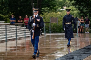 A soldier wearing the Army ceremonial dress uniform (dark jacket, lighter blue pants with gold stripe down the leg) is carrying a rifle in both hands in front of his body with the bayonet pointing up to the sky. There is another soldier dressed similarly but with a raincoat on following him a few feet back. There is a back mat on the ground nearby, and they are both walking on the marble walkway. There are few people in the background watching. The ground is wet.