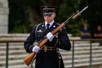 A male soldier wearing sunglasses and dressed in Army ceremonial dress uniform (dark jacket with gold buttons and badges, multicolored ribbons, and light blue shoulder braid) is holding a rifle at a 45 degree angle in both hands in front of his body while either standing or marching near some light colored steps.
