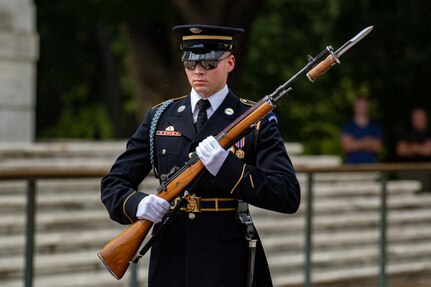 A male soldier wearing sunglasses and dressed in Army ceremonial dress uniform (dark jacket with gold buttons and badges, multicolored ribbons, and light blue shoulder braid) is holding a rifle at a 45 degree angle in both hands in front of his body while either standing or marching near some light colored steps.