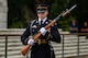 A male soldier wearing sunglasses and dressed in Army ceremonial dress uniform (dark jacket with gold buttons and badges, multicolored ribbons, and light blue shoulder braid) is holding a rifle at a 45 degree angle in both hands in front of his body while either standing or marching near some light colored steps.