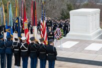 Members of the armed forces in various types of ceremonial uniforms are holding flags (US and other service flags) in front of the white marble Tomb of the Unknown Soldier (large, rectangular shaped). In front of the tomb is a multicolored wreath on a stand. Other service members are standing in rows in a formation near the tomb.