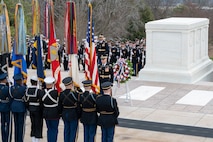 Members of the armed forces in various types of ceremonial uniforms are holding flags (US and other service flags) in front of the white marble Tomb of the Unknown Soldier (large, rectangular shaped). In front of the tomb is a multicolored wreath on a stand. Other service members are standing in rows in a formation near the tomb.