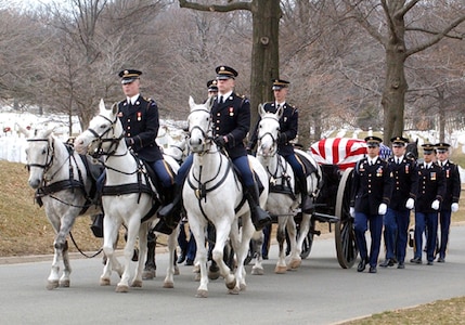 Soldiers in dark ceremonial uniforms are riding on white horses that are pulling a dark caisson with large wooden wheels that has a U.S. flag-draped casket on it. There are other soldiers in dark ceremonial uniforms marching alongside the caisson.