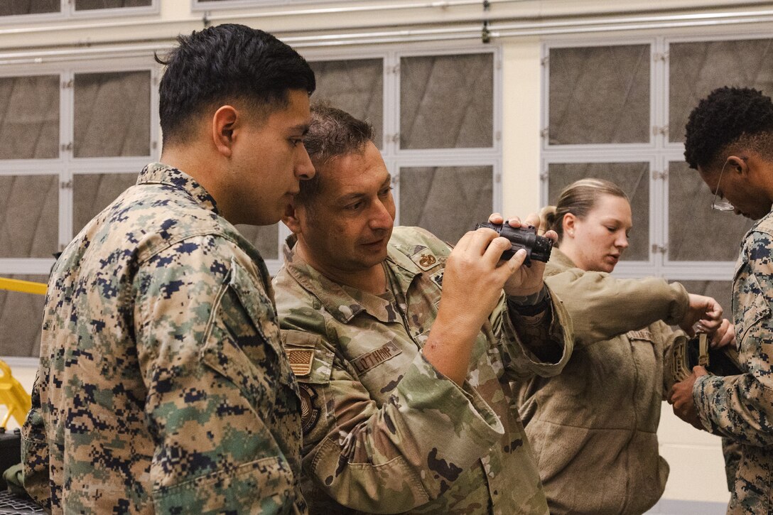 U.S. Air Force Tech. Sgt. Matthew Lothrop, an air transportation specialist with the U.S. Air Force 36th Contingency Response Squadron, 36th Contingency Response Group, center, shows U.S. Marine Corps Lance Cpl. Abel Gamez, a distribution management specialist with the Marine Corps Air Station Iwakuni Distribution Management Office, left, how to operate Gen III AN/AVS-9 night vision goggles during a training exercise at Marine Corps Air Station Iwakuni, Japan, Dec. 13, 2023.  The U.S. Air Force 36th Contingency Response Group conducted joint training that focused on equipment and process interoperability in a contested logistics environment with the MCAS Iwakuni Headquarters and Headquarters Squadron Logistics Department and members of the Japan Air Self-Defense Force.  (U.S. Marine Corps photo by Cpl. Darien Wright)