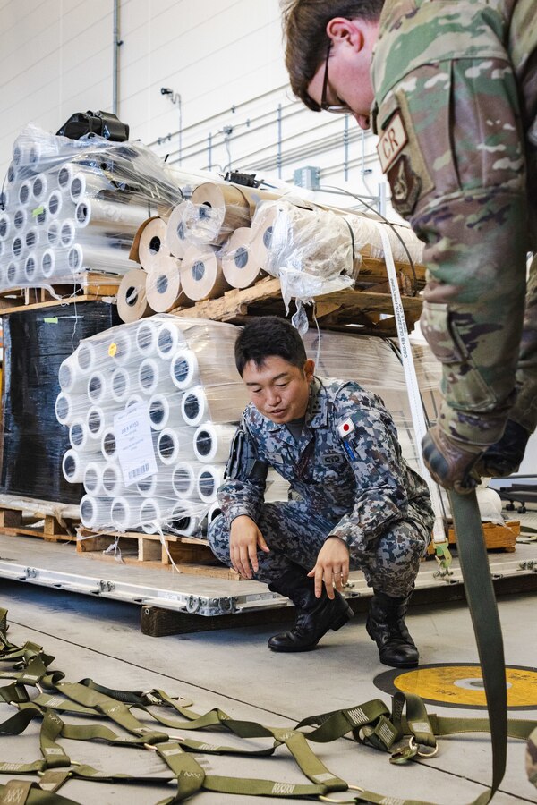 Japan Air Self-Defense Force Capt. Atsushi Kanda, an air traffic controller with Air Traffic Control Service Group, Air Support Command, left, watches as U.S. Air Force Senior Airman Dalton Calhoun, an aerial port journeyman with the U.S. Air Force 36th Contingency Response Squadron, 36th Contingency Response Group, demonstrates how to secure cargo during a training exercise at Marine Corps Air Station Iwakuni, Japan, Dec. 12, 2023. The U.S. Air Force 36th Contingency Response Group conducted joint training that focused on equipment and process interoperability in a contested logistics environment with the MCAS Iwakuni Headquarters and Headquarters Squadron Logistics Department and members of the Japan Air Self-Defense Force.  (U.S. Marine Corps photo by Cpl. Darien Wright)