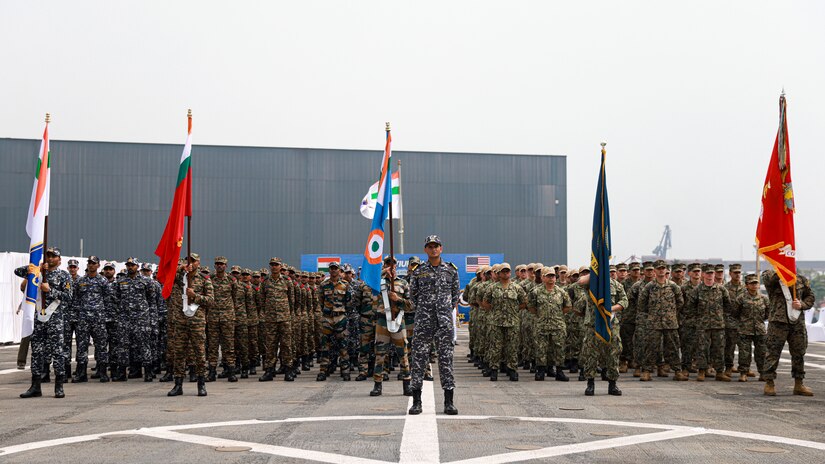 U.S. and Indian service members stand in formation holding flags aboard  a ship.