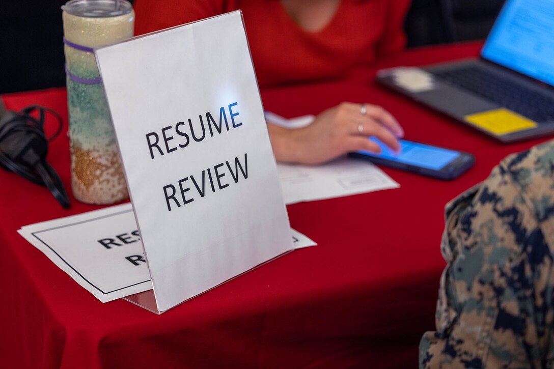 A "RESUME REVIEW" sign sits on a table with a service member on one side and a civilian using a phone and laptop on the other.