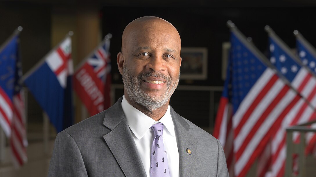 Head and shoulders shot of a black man in a suit in front of flags